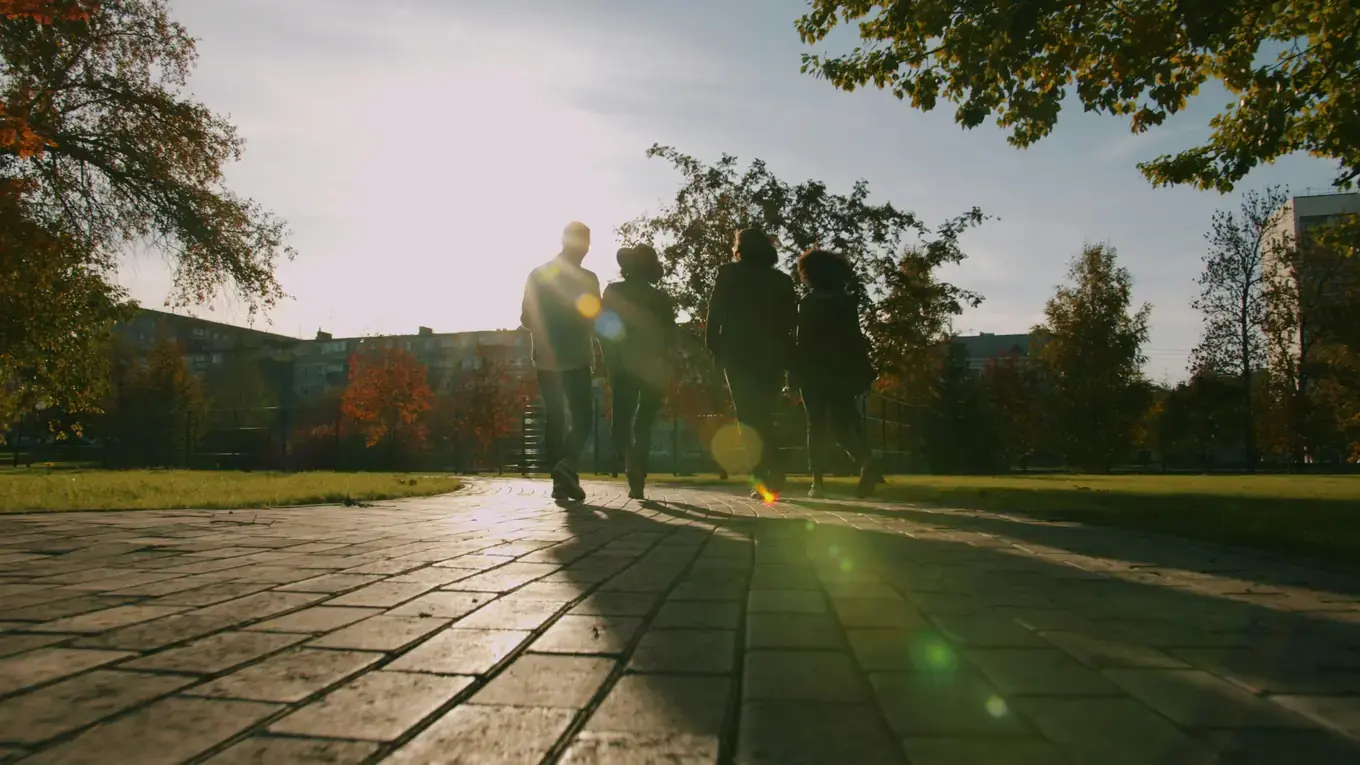 Four people, seen from behind as dark silhouettes, walk side by side along a paved park path into bright sunlight. Trees with orange leaves and blocks of flats stand in the background.