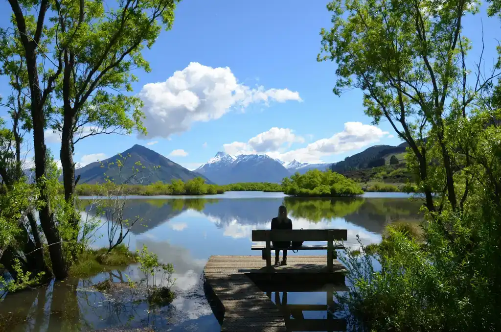 A person with long fair hair sits alone on a bench at the end of a wooden boardwalk. Still water reflects clouds, trees and snow-capped mountains.