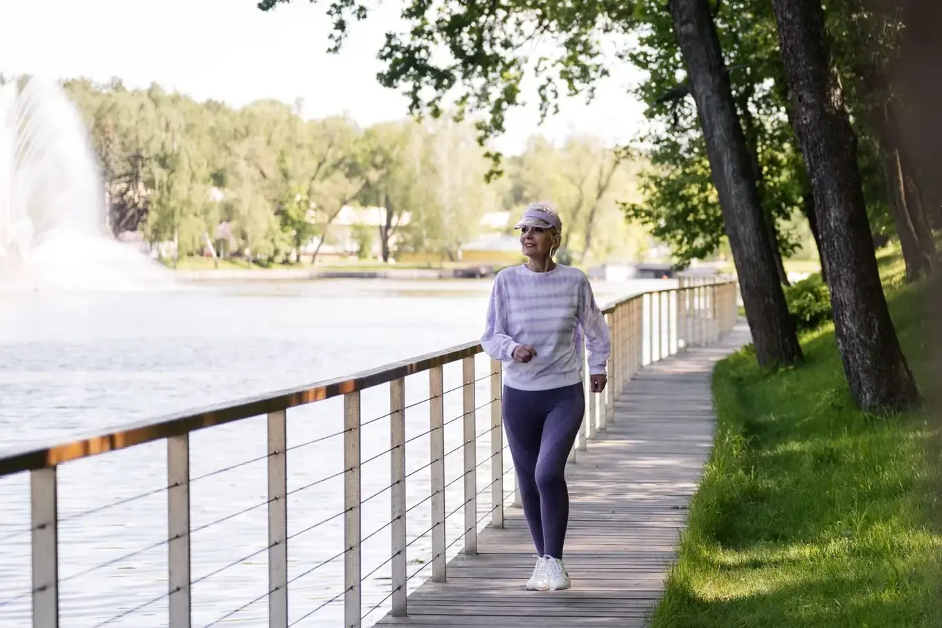 A smiling older woman in a lilac visor, a striped lilac sweatshirt, purple leggings and white trainers walks along a wooden boardwalk. A railing separates the boardwalk from a lake with a fountain, and trees stand on the grass to the right.