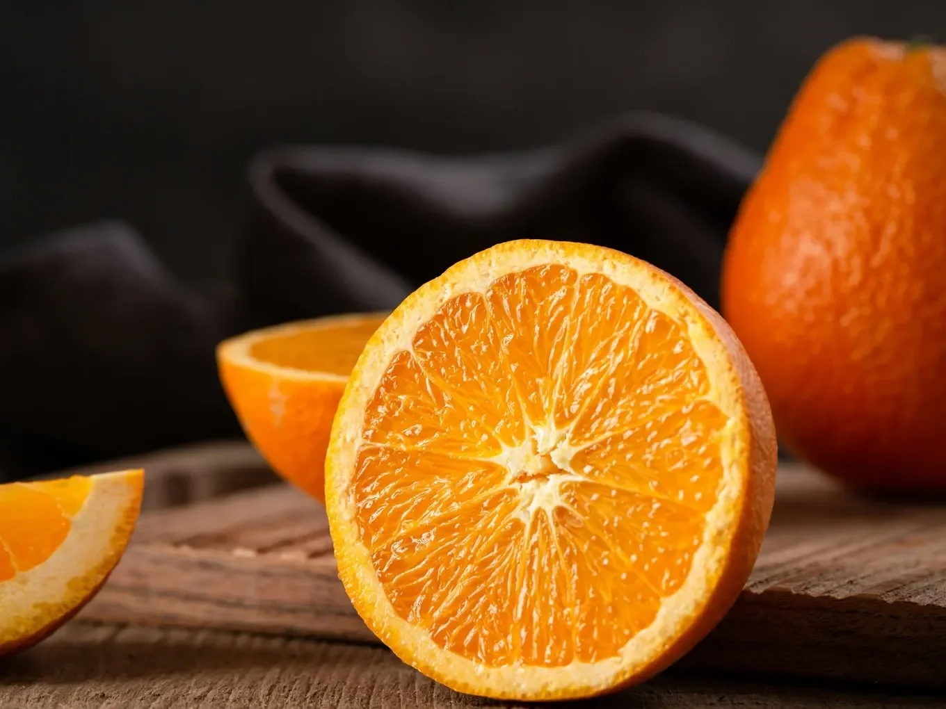 A halved orange stands cut side forward on a wooden board, with another half and a wedge beside it and a whole orange behind, against a dark cloth.
