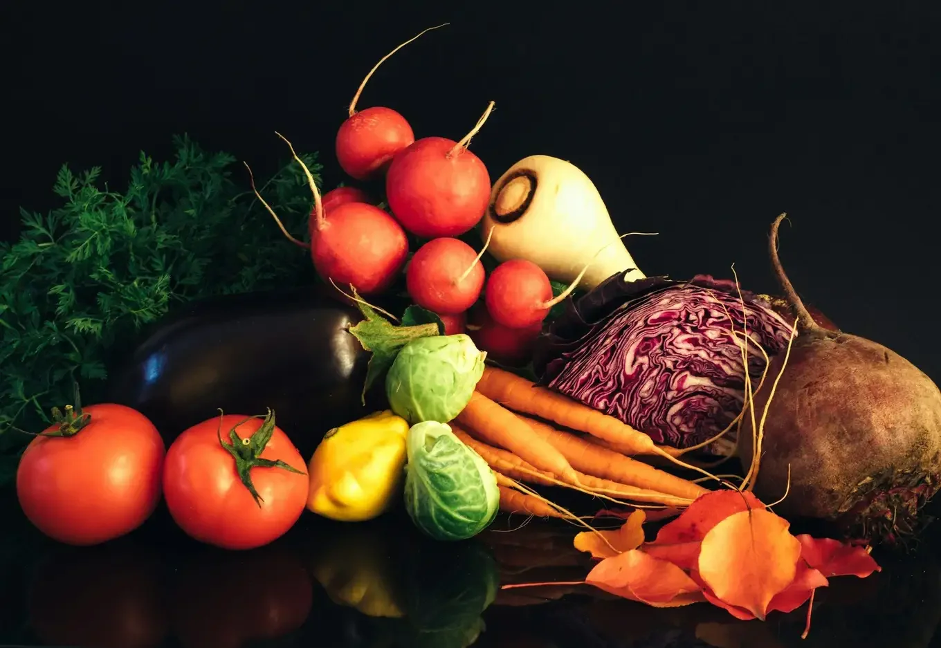 Fresh vegetables heaped on a black surface: tomatoes, an aubergine, radishes, a parsnip, red cabbage, carrots, Brussels sprouts, a beetroot and a yellow squash, with carrot tops and orange autumn leaves.