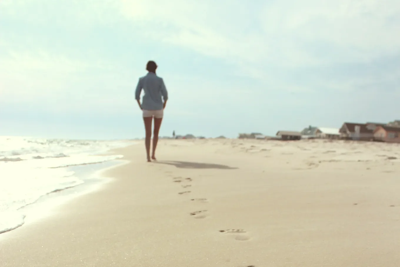 A person in a light blue shirt and white shorts heads away from the camera along a sandy beach. Footprints trail back through the sand, with waves on the left and low houses on the right.