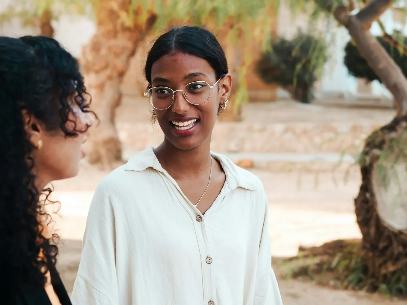 Two people face each other outdoors under trees. One, in glasses and a cream shirt, smiles at the other, who has long curly hair and dark clothes.