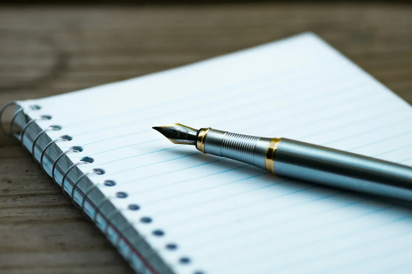 A silver fountain pen with gold trim rests on an empty page ruled in pale blue. The spiral binding lies on a weathered wooden table.